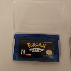 May include: A translucent blue Game Boy Advance video game cartridge for Pokémon Sapphire Version, housed in a clear plastic protective case. The label features the game title, the ESRB Everyone rating, the Nintendo logo, and code AGB-AXPE-USA.