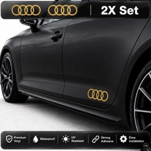 May include: A 2X set of gold-coloured Audi four-ring logo decals applied to the side of a matte black car. The decals feature the iconic interlocking ring design. The image includes text labels for premium vinyl, waterproof, UV resistant, strong adhesive, and easy installation.