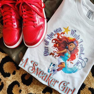 May include: White t-shirt with a graphic of a mermaid with red hair and a blue tail. The text "Wish I could stay out of the sea" is written in a purple font. The text "A Sneaker Girl" is written in a glittery red font.