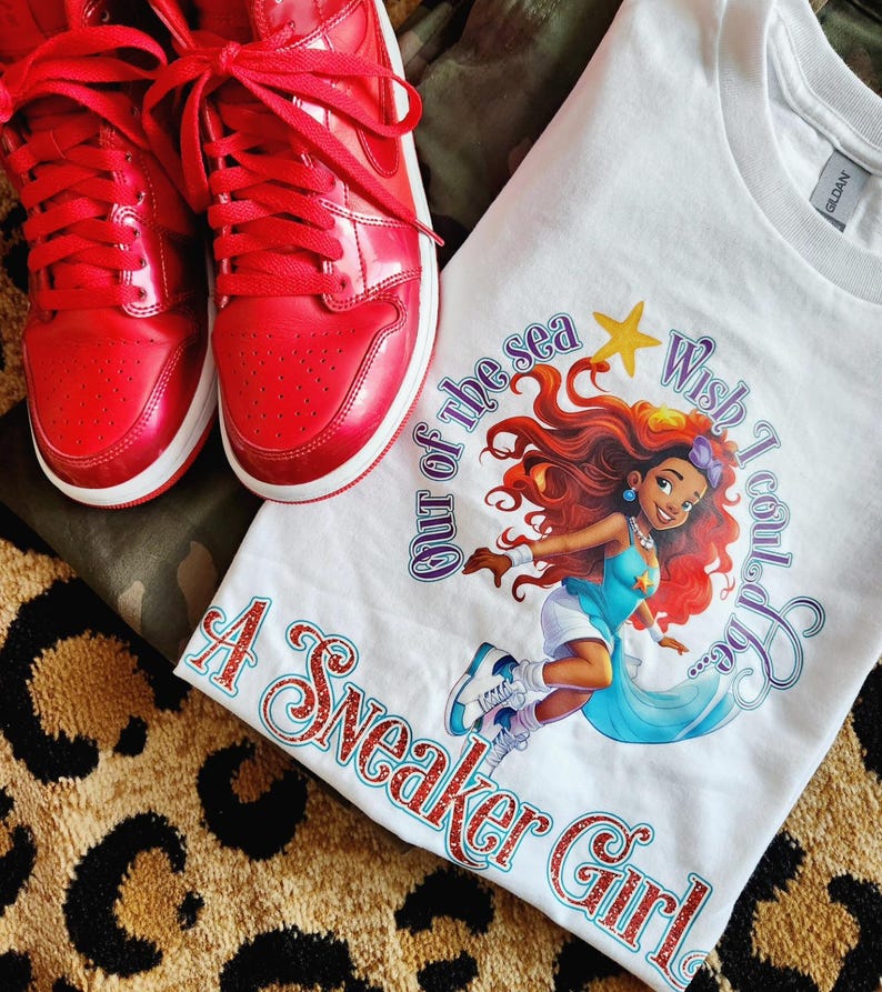 May include: White t-shirt with a graphic of a mermaid with red hair and a blue tail. The text "Wish I could stay out of the sea" is written in a purple font. The text "A Sneaker Girl" is written in a glittery red font.