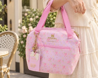 Pode incluir: Uma bolsa térmica rosa com padrão floral, logótipos dourados Stanley e LoveShackFancy e um bolso frontal com fecho. A bolsa inclui pegas de tecido rosa e um pendente decorativo em tom dourado com um laço branco e uma etiqueta.