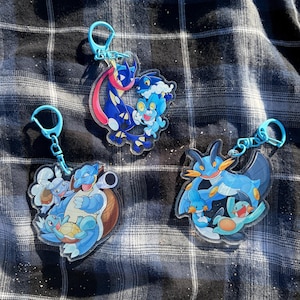 Könnte beinhalten: Drei durchsichtige Acryl-Schlüsselanhänger mit farbenfrohen Comic-Kreaturen-Illustrationen. Jeder Anhänger hat einen hellblauen Kunststoff-Karabinerverschluss. Die Designs zeigen verschiedene blau getönte Figuren in dynamischen Posen.