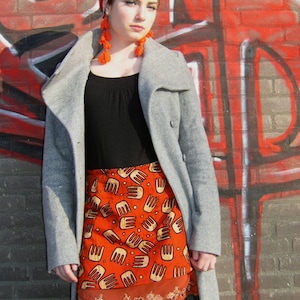 May include: A woman wearing a gray coat with a black top and a red and orange patterned skirt. She is wearing a headscarf and orange tassel earrings.