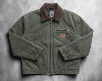 Puede incluir: Una chaqueta de lona verde oliva con cuello de pana marrón, cremallera frontal completa y un bolsillo en el pecho con cremallera. Un parche cuadrado de la marca Carhartt es visible en el pecho. El forro interior muestra un patrón de cuadros.