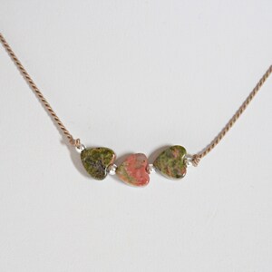 May include: A necklace with three heart-shaped stones. The stones are green, pink, and green. The necklace is made of brown cord.