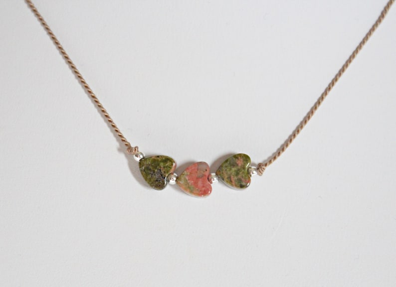 May include: A necklace with three heart-shaped stones. The stones are green, pink, and green. The necklace is made of brown cord.