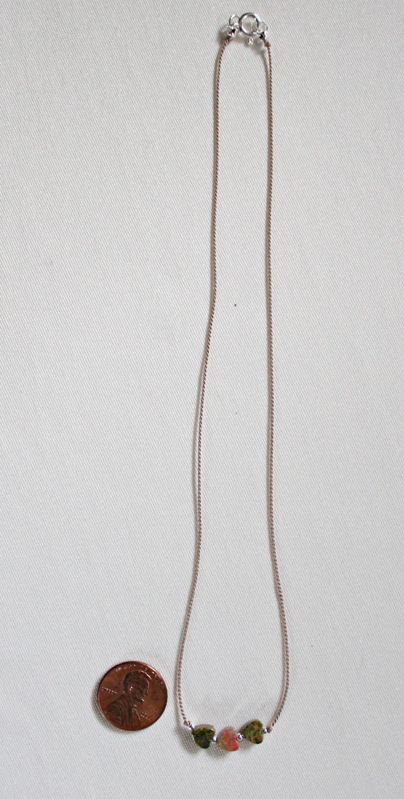 May include: A silver chain necklace with three heart-shaped stones. The stones are green, pink, and green. The necklace is about the size of a US penny.