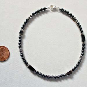 Op de afbeelding: Een donkergrijze enkelband met zilverkleurige accenten en rechthoekige kralen. De enkelband wordt weergegeven op een wit oppervlak naast een cent ter referentie. De enkelband is ongeveer 23 cm lang.