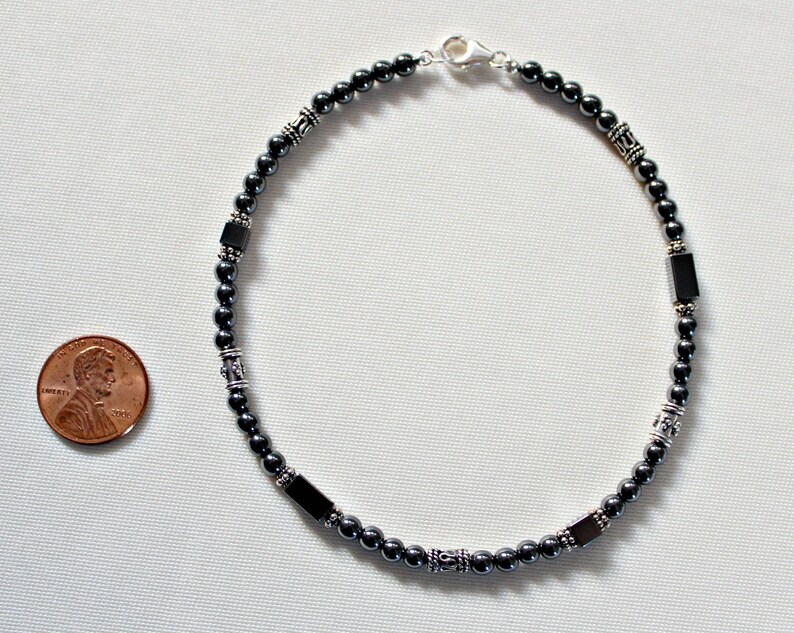 Op de afbeelding: Een donkergrijze enkelband met zilverkleurige accenten en rechthoekige kralen. De enkelband wordt weergegeven op een wit oppervlak naast een cent ter referentie. De enkelband is ongeveer 23 cm lang.