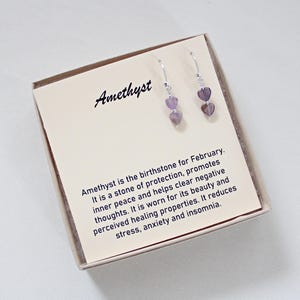 May include: Amethyst heart earrings on a card. The card reads "Amethyst" and describes the stone's properties. The earrings have two heart-shaped purple stones. The earrings are displayed in a small, light-colored box.