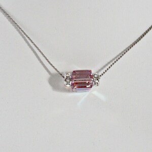 May include: A silver chain necklace with a pink crystal pendant. The pendant is a rectangular prism with clear crystal accents on either side.