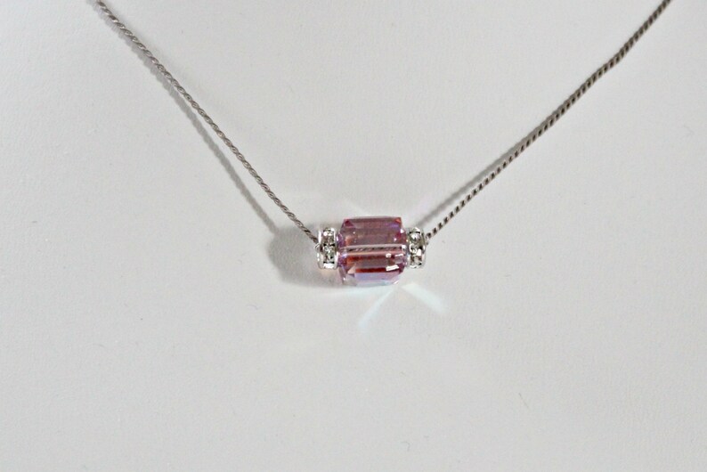May include: A silver chain necklace with a pink crystal pendant. The pendant is a rectangular prism with clear crystal accents on either side.