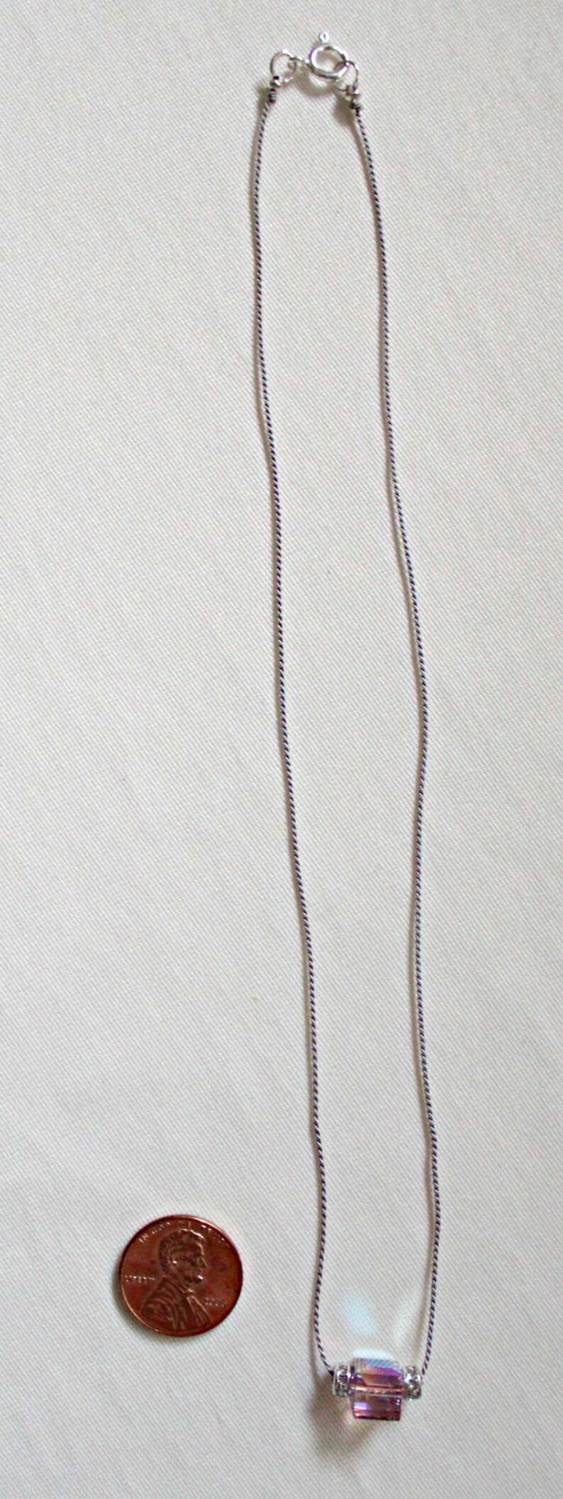May include: A silver chain necklace with a small, iridescent, rectangular crystal pendant. The pendant is surrounded by two silver rings with small clear crystals. The necklace is shown next to a US penny for size comparison.