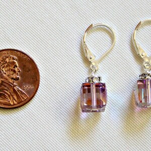 May include: A pair of silver earrings with a dangling square-shaped crystal. The crystal is a light purple color with a shimmery finish. The earrings are about the size of a US penny.