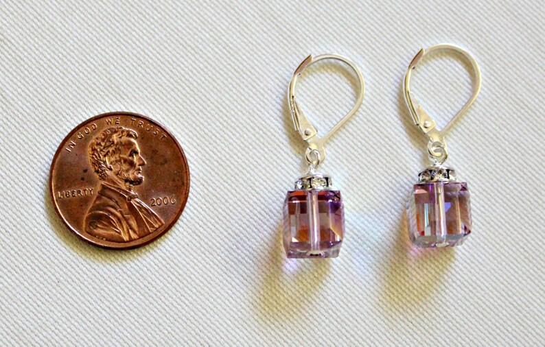 May include: A pair of silver earrings with a dangling square-shaped crystal. The crystal is a light purple color with a shimmery finish. The earrings are about the size of a US penny.