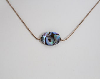 May include: Abalone shell pendant necklace. The oval-shaped pendant features iridescent blues, greens, and purples. The pendant is strung on a delicate, light brown cord, creating a minimalist design. The necklace is a piece of jewelry.