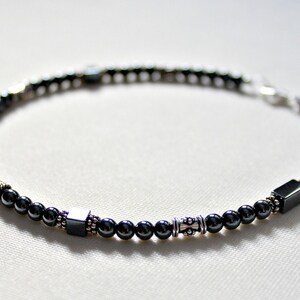 Op de afbeelding: Een kralenarmband met kleine, donkergrijze kralen afgewisseld met zilverkleurige accenten en vierkante kralen. De armband is geregen op een flexibel koord en heeft een zilveren sluiting. De armband is ongeveer 19 cm lang.