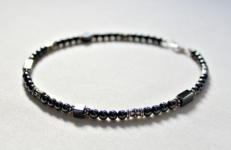 Op de afbeelding: Een kralenarmband met kleine, donkergrijze kralen afgewisseld met zilverkleurige accenten en vierkante kralen. De armband is geregen op een flexibel koord en heeft een zilveren sluiting. De armband is ongeveer 19 cm lang.