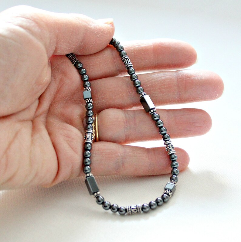 Op de afbeelding: Een donkergrijze kralenketting met zilverkleurige accenten. De ketting heeft kleine, ronde kralen afgewisseld met vierkante en cilindrische zilverkleurige elementen. De ketting wordt in een hand gehouden tegen een witte achtergrond.