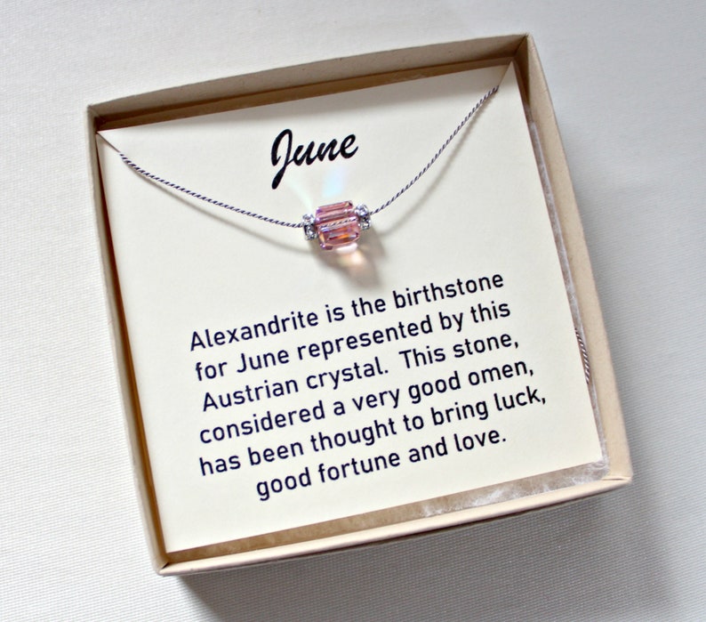 May include: A small box with a white card inside. The card has the word "June" printed on it and a description of Alexandrite, the birthstone for June. A delicate necklace with a pink crystal bead is displayed on the card.
