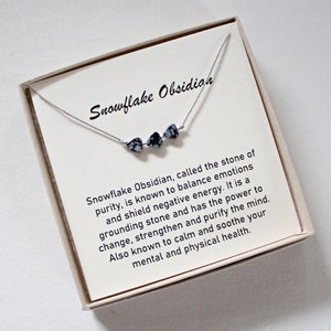 May include: A delicate necklace featuring three snowflake obsidian beads on a thin silver chain. The necklace is presented on a card inside a beige box. The card reads "Snowflake Obsidian" and describes the stone's properties.