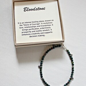 May include: A green bloodstone bracelet with silver beads and charms. The bracelet is in a small box with a description of the stone's properties. "Bloodstone It is an intense healing stone, known as the "Stone of Courage". It revitalizes love, relationships and friendships. It brings purification, orderliness, prosperity and instills wisdom. It enhances creativity and supports decision making."