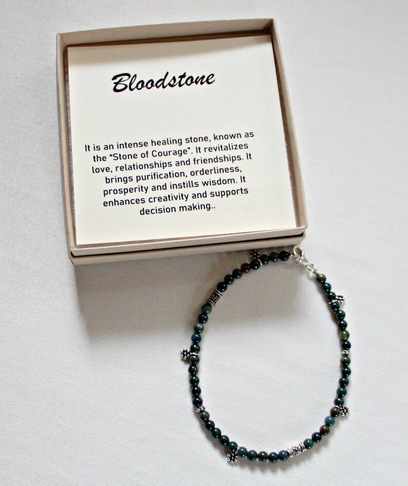 May include: A green bloodstone bracelet with silver beads and charms. The bracelet is in a small box with a description of the stone's properties. "Bloodstone It is an intense healing stone, known as the "Stone of Courage". It revitalizes love, relationships and friendships. It brings purification, orderliness, prosperity and instills wisdom. It enhances creativity and supports decision making."
