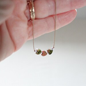May include: A delicate gold chain necklace with three small heart-shaped pendants. The pendants are a mottled green and brown color.