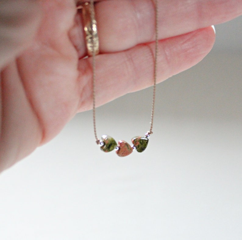 May include: A delicate gold chain necklace with three small heart-shaped pendants. The pendants are a mottled green and brown color.