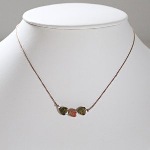 May include: A gold chain necklace with three heart-shaped pendants. The pendants are made of green, pink, and brown stone.