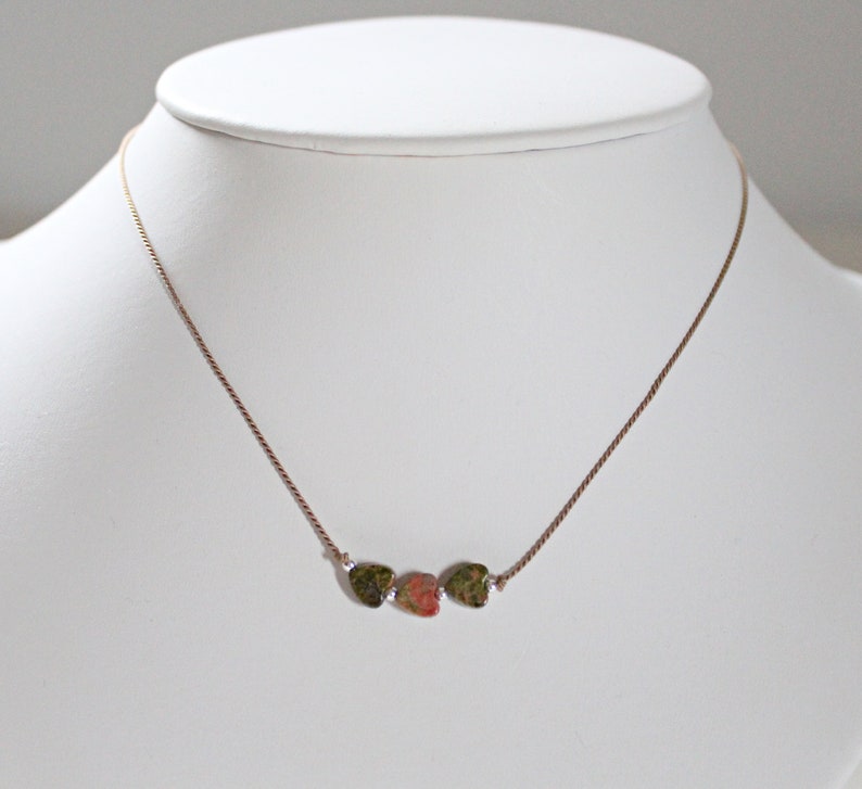 May include: A gold chain necklace with three heart-shaped pendants. The pendants are made of green, pink, and brown stone.