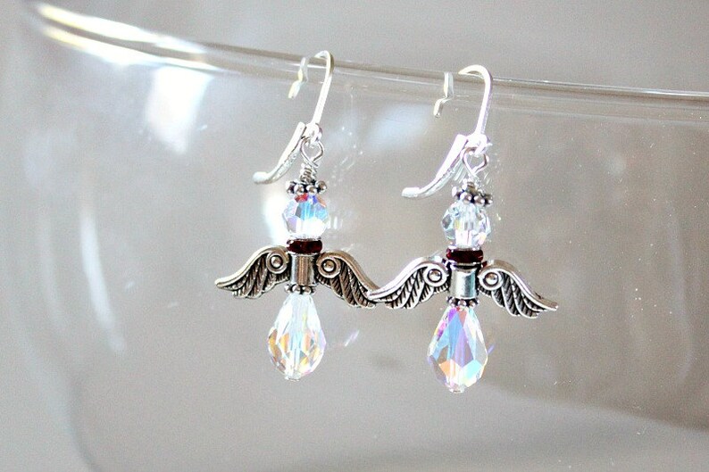 May include: Silver dangle earrings with a crystal teardrop and a silver angel wing charm. The angel wings have a dark red accent.