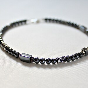 Op de afbeelding: Een donkergrijze kralenarmband met een mix van kleine ronde kralen en grotere vierkante en rechthoekige kralen. De armband heeft een zilveren sluiting en decoratieve zilveren accenten. De kralen zijn geregen op een flexibel koord.