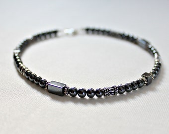 May include: A dark gray beaded bracelet with a mix of small round beads and larger square and rectangular beads. The bracelet has a silver clasp and decorative silver accents. The beads are strung on a flexible cord.