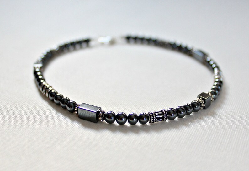 Op de afbeelding: Een donkergrijze kralenarmband met een mix van kleine ronde kralen en grotere vierkante en rechthoekige kralen. De armband heeft een zilveren sluiting en decoratieve zilveren accenten. De kralen zijn geregen op een flexibel koord.
