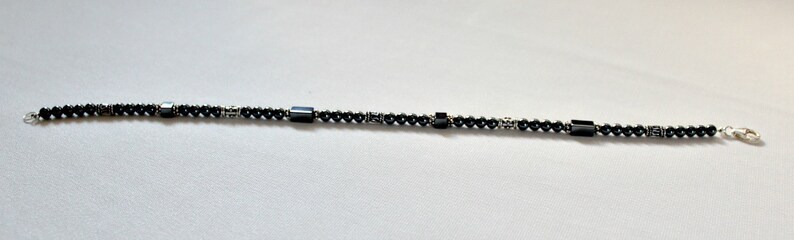 Op de afbeelding: Een zwart kralenarmband met een zilveren sluiting. De armband heeft kleine, ronde zwarte kralen afgewisseld met rechthoekige en cilindrische zwarte kralen. De sluiting en verbindingsstukken zijn zilverkleurig.