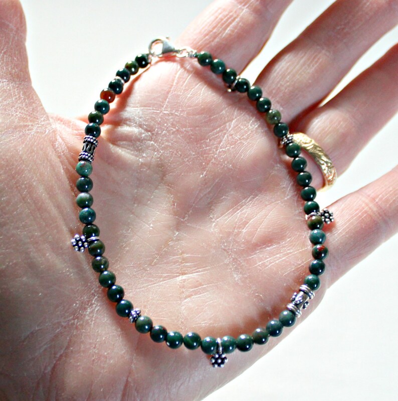 May include: A green and silver beaded bracelet with silver flower charms. The bracelet is made with small, round beads and has a silver clasp.