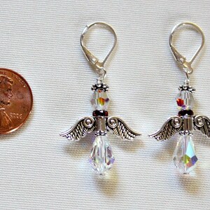 May include: A pair of silver angel wing earrings with clear crystal beads and a red bead accent. The earrings are about the size of a US penny.