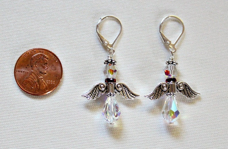 May include: A pair of silver angel wing earrings with clear crystal beads and a red bead accent. The earrings are about the size of a US penny.
