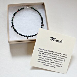 May include: A green bloodstone necklace with silver charms in a white box. The box has a card with the text "March" and information about the birthstone for March.