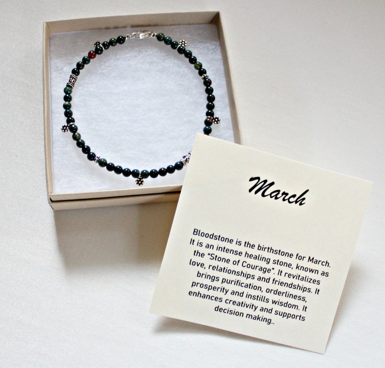 May include: A green bloodstone necklace with silver charms in a white box. The box has a card with the text "March" and information about the birthstone for March.