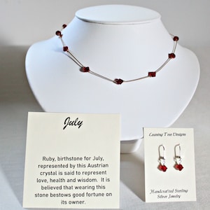 May include: A red ruby necklace and earrings set on a white display stand. The necklace is made of a thin gold chain with red ruby beads. The earrings are silver with red ruby stones. The card reads "July, Ruby, birthstone for July, represented by this Austrian crystal is said to represent love, health and wisdom. It is believed that wearing this stone bestows good fortune on its owner." and "Ceaning Tree Designs, Handcrafted Sterling Silver Jewelry".