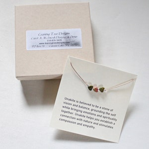 May include: A small, delicate necklace with three green, brown, and pink Unakite stones. The necklace is on a white card with text that reads: "Unakite is believed to be a stone of vision and balance, grounding the self while bringing emotions and spirituality together. Unakite helps you establish a connection with nature and stimulates compassion and empathy."
