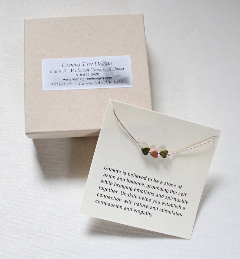 May include: A small, delicate necklace with three green, brown, and pink Unakite stones. The necklace is on a white card with text that reads: "Unakite is believed to be a stone of vision and balance, grounding the self while bringing emotions and spirituality together. Unakite helps you establish a connection with nature and stimulates compassion and empathy."