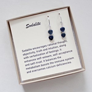 May include: A pair of silver earrings with blue sodalite heart-shaped stones. The earrings are in a white box with a description of the stone's properties. Sodalite encourages rational thought, objectivity, truth and intuition, along with verbalization of feelings. It enhances self-esteem, self-acceptance and self-trust. It balances the metabolism boosts the immune system and overcomes calcium deficiencies.