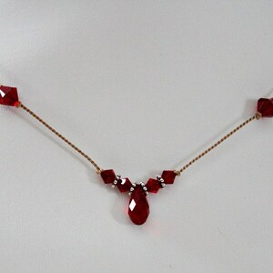 May include: A delicate necklace featuring a series of faceted, deep red crystals. The crystals are interspersed along a thin, gold-colored chain, with a teardrop-shaped crystal pendant at the center. The necklace is designed for a refined look.