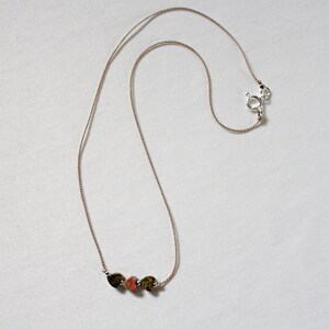 May include: A delicate necklace with a brown cord and three small heart-shaped stones. The stones are a mix of green, brown, and pink.