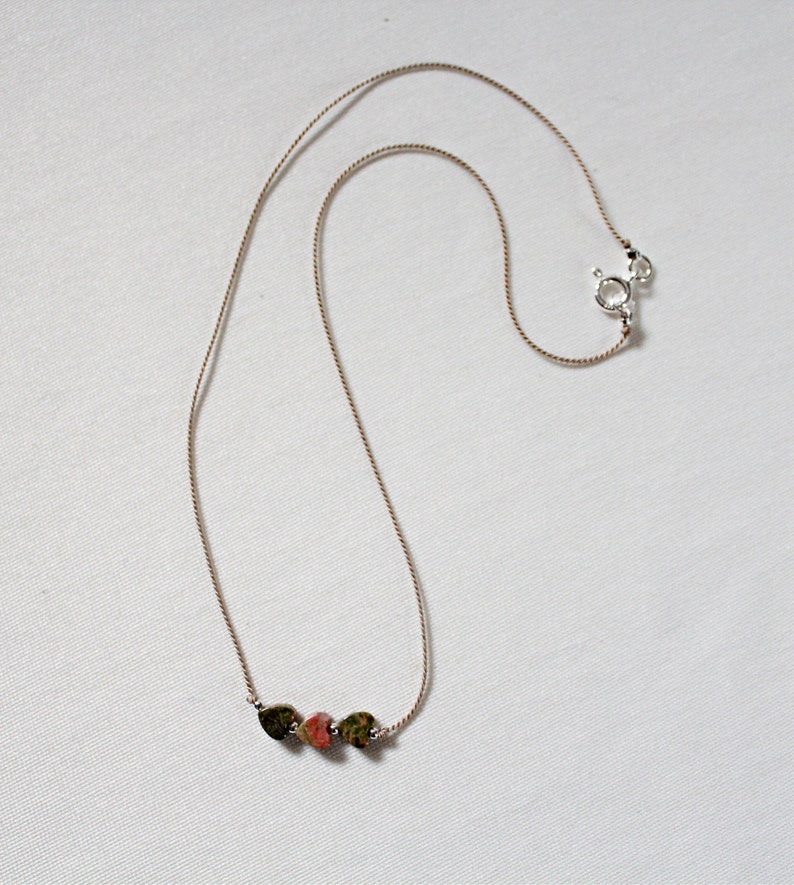 May include: A delicate necklace with a brown cord and three small heart-shaped stones. The stones are a mix of green, brown, and pink.