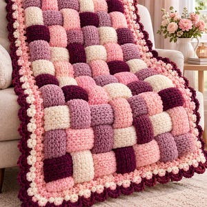 Puede incluir: Una manta de ganchillo gruesa con un patrón de tejido de cesta en tonos crema, rosa claro, malva y burdeos oscuro. La manta está rematada con un borde decorativo festoneado en hilos de colores crema y burdeos oscuro alternados.