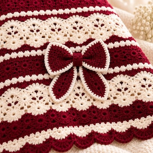 May include: A crochet blanket featuring alternating horizontal stripes of deep burgundy and cream yarn. The design incorporates intricate floral lace patterns and a prominent, centered burgundy crochet bow with a cream-colored border trim.